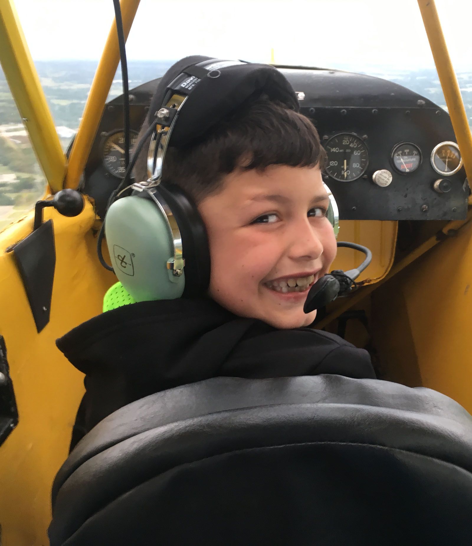 young man wearing headset in front seat of piper cub, turning around and smiling
